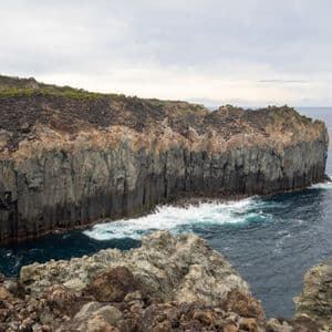 Une haute falaise de basalte en colonnes rencontre la mer sombre, avec des vagues blanches s'écrasant contre sa base rocheuse.