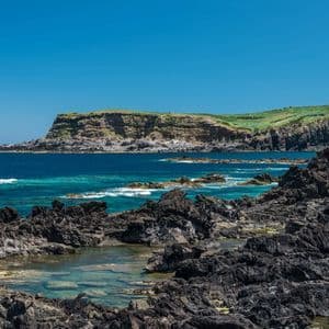 Un littoral rocheux et volcanique avec des piscines naturelles, une mer turquoise et des falaises vertes lointaines sous un ciel bleu clair.