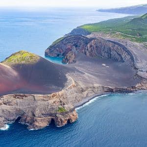 Vue aérienne d'une péninsule volcanique avec terre rouge et noire, rencontrant des falaises escarpées et l'océan d'un bleu profond.