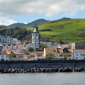 Une ville côtière avec des bâtiments colorés et un clocher d'église sombre se dresse derrière une digue en pierre, avec une grande colline verte en arrière-plan.
