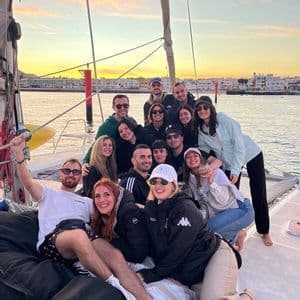 Un gruppo WeRoad sorride per una foto sul ponte di una barca a vela al tramonto, con una città costiera sullo sfondo.
