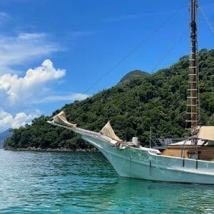 La prua di una barca a vela bianca galleggia su acque tranquille e turchesi vicino a un'isola lussureggiante e alberata sotto un cielo azzurro.