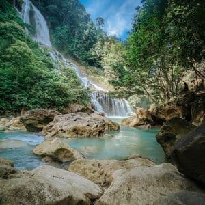 Un'alta cascata scende da un fianco boscoso in una pozza rocciosa dalle acque turchesi, con una persona seduta su una roccia lì vicino.