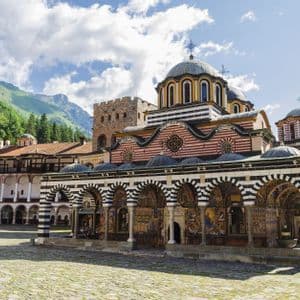 Un monastero ornato con archi a strisce e cupole si erge in un cortile acciottolato, con montagne boscose sullo sfondo.