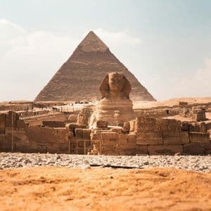 Una vista desde el suelo de la Gran Esfinge con una gran pirámide al fondo, rodeada de antiguas ruinas de piedra bajo un cielo brillante.