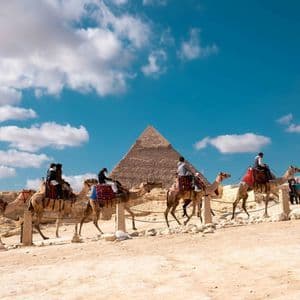 Un viaje en grupo de WeRoad montando en una fila de camellos en el desierto, con una gran pirámide al fondo bajo un cielo azul con nubes.