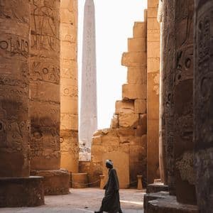 Una persona con atuendo tradicional camina junto a grandes columnas de piedra con jeroglíficos, con un obelisco al fondo.