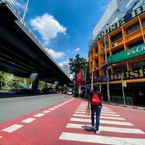 Una persona con una mochila roja camina por un paso de cebra rojo y blanco a lo largo de una calle de la ciudad junto a un paso elevado.