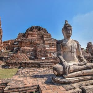 Una estatua de Buda de piedra se sienta en pose meditativa frente a antiguas ruinas de templo de ladrillo rojo bajo un cielo azul claro.