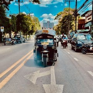 Un taxi tuk-tuk azul circula por una concurrida calle de la ciudad, pasando sobre una gran flecha pintada, con árboles y cables eléctricos en lo alto.