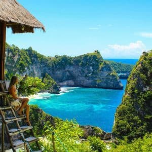 Una mujer con un vestido floral se sienta en una escalera de madera junto a una casa en el árbol, con vistas a una bahía tropical con acantilados y agua turquesa.