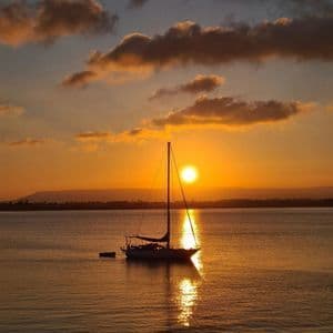 Una barca a vela galleggia sull'acqua calma mentre il sole tramonta, proiettando un riflesso dorato sulla superficie.