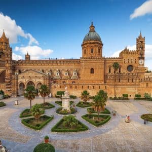 Una cattedrale in pietra, riccamente decorata, con una grande cupola si affaccia su una piazza pavimentata con giardini geometrici e palme sotto un cielo blu e nuvoloso.