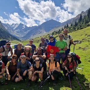 Un groupe WeRoad en voyage avec sacs à dos et bâtons de trekking pose pour une photo dans une verte vallée de montagne, l'un d'eux brandissant un drapeau du Kirghizistan.
