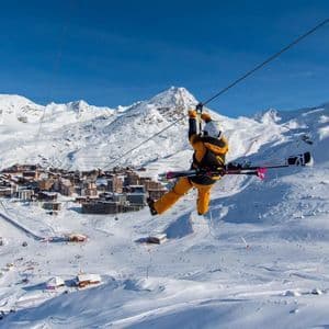 A person wearing ski gear glides down a zip line with skis on their back, high above a snowy mountain village.