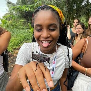 Una donna durante un viaggio di gruppo WeRoad sorride e fa la linguaccia mentre tiene in mano un grosso insetto nero in un ambiente tropicale.