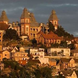 A dense cluster of historic buildings with towers sits on a hillside, illuminated by the warm light of sunset against a dark sky.