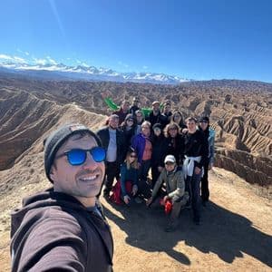 Un hombre se toma una selfie con un viaje en grupo de WeRoad en una cresta con vistas a un cañón, con montañas nevadas bajo un cielo soleado.