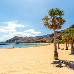 Palmen werfen Schatten auf einen goldenen Sandstrand, der an eine ruhige blaue Bucht grenzt, mit schroffen Bergen im Hintergrund unter einem sonnigen Himmel.