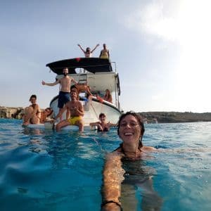 Una mujer se toma una selfie sonriendo mientras nada en el mar con su grupo de viaje WeRoad relajándose en un barco al fondo.