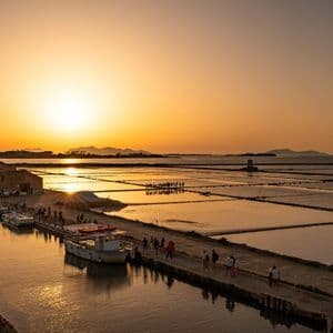 Un viaje en grupo de WeRoad observa el atardecer dorado sobre las salinas junto a un canal con barcos amarrados y un molino de viento tradicional.