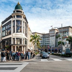 Peatones esperando en un cruce peatonal frente a un edificio esquinero ornamentado en una calle soleada de la ciudad con palmeras.