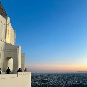 Varias personas están en el balcón de un edificio blanco, contemplando una extensa ciudad al atardecer.
