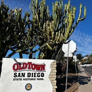 El letrero blanco de entrada del Parque Histórico Estatal Old Town San Diego se alza frente a un gran cactus bajo un cielo azul.