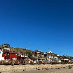Una hilera de casas coloridas se asienta en una ladera verde sobre una playa de arena bajo un cielo azul claro.