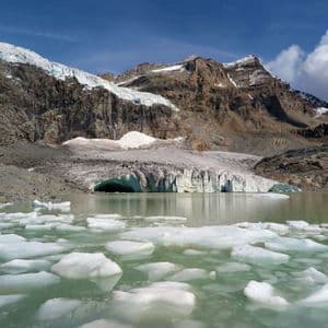 Un lago glaciale con iceberg galleggianti si trova di fronte a un enorme ghiacciaio incastonato tra montagne rocciose e coperte di neve.