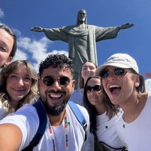A WeRoad group trip takes a smiling selfie in front of a large statue of a man with outstretched arms.