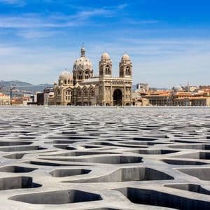 Una vista de una gran catedral a través de una moderna terraza de hormigón estampado, con un paisaje urbano de fondo bajo un cielo azul.