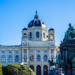 Un bâtiment historique orné d'un grand dôme se dresse derrière un monument imposant avec plusieurs statues sous un ciel bleu clair.