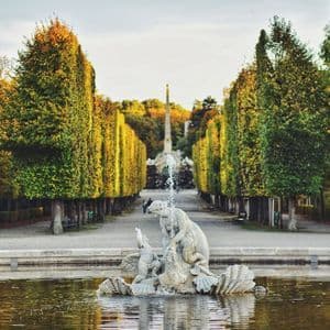 Une fontaine en pierre dans un étang, au début d'une longue allée bordée de grands arbres taillés au feuillage d'automne.