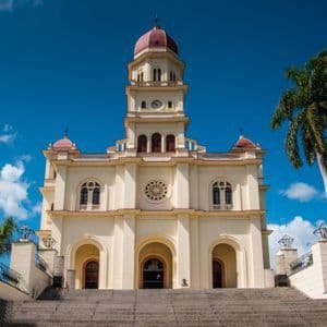 Vista en contrapicado de una gran iglesia de color crema con torre de cúpula roja, desde la base de una amplia escalera de piedra.