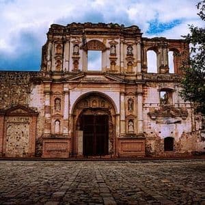La facciata logora di una chiesa in pietra in rovina si erge dietro una piazza acciottolata sotto un cielo nuvoloso.
