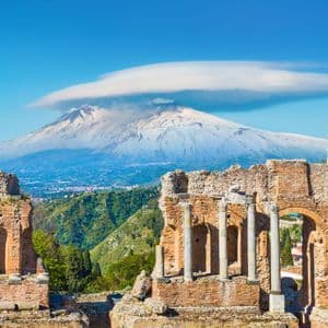 Ancient brick theater ruins frame a distant, snow-capped volcano under a clear blue sky with a single cloud.