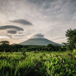Un volcan avec un nuage lenticulaire au-dessus de son sommet, s'élevant au-dessus d'un paysage verdoyant de champs et de forêt au coucher du soleil.