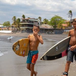 Deux jeunes garçons avec des planches de surf marchent le long du rivage sur une plage de sable avec des palmiers en arrière-plan.