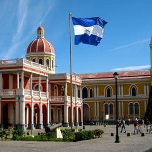 Le drapeau du Nicaragua flotte au-dessus d'une place coloniale ensoleillée avec des bâtiments rouges et jaunes, des dômes et un canon.
