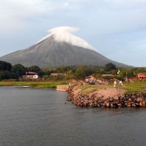 Un volcan avec un nuage lenticulaire sur son sommet surplombe un plan d'eau avec un bateau et une jetée rocheuse au premier plan.