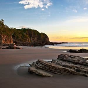 Une personne assise sur un rocher sur une plage de sable, avec des falaises couvertes d'arbres et l'océan visibles lors d'un coucher de soleil doré.