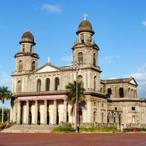 Une grande cathédrale de pierre avec deux clochers à dôme et une entrée à colonnes, vue depuis une place en briques rouges.