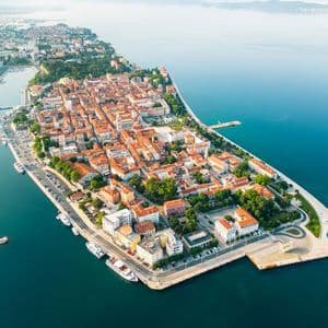 Una vista aerea di una città costiera su una penisola, con edifici dai tetti in terracotta circondati dal mare.