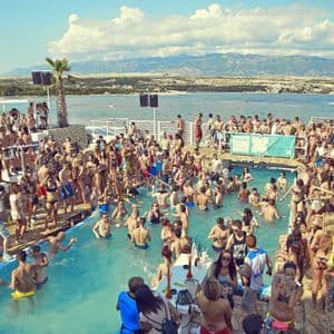 Un viaggio di gruppo WeRoad a una festa in piscina in un affollato beach club, con il mare e le montagne distanti sullo sfondo.