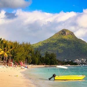 Un bateau à moteur jaune repose dans l'eau turquoise d'une plage de sable bordée de palmiers, avec une grande montagne verte en arrière-plan.