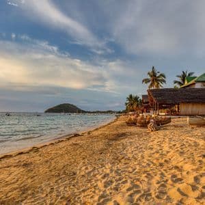 Ein Sandstrand mit Strohdachhütten und Booten am Ufer während eines goldenen Sonnenuntergangs.