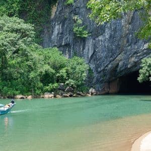 Una barca turistica blu con persone naviga su un fiume turchese, dirigendosi verso una grande apertura di grotta alla base di una montagna boscosa.