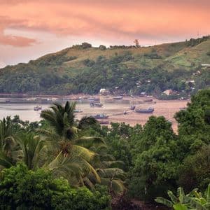 Blick aus der Vogelperspektive über üppige Baumwipfel auf eine Bucht mit vielen Booten bei Ebbe, dahinter ein großer grüner Hügel unter rosafarbenem Abendhimmel.
