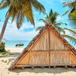 Eine dreieckige Holzhütte mit Strohdach steht an einem weißen Sandstrand unter Palmen, mit einem Boot auf dem türkisfarbenen Wasser.